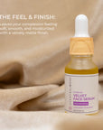 Scentral Zone 0.5 fl oz Velvet Face Serum, a yellow rejuvenating serum with cucumber and seaweed, in a glass dropper bottle with a wooden collar and white dropper top. Product is displayed with a light brown fabric backdrop and test explaining the feel and finish of the product after applying.