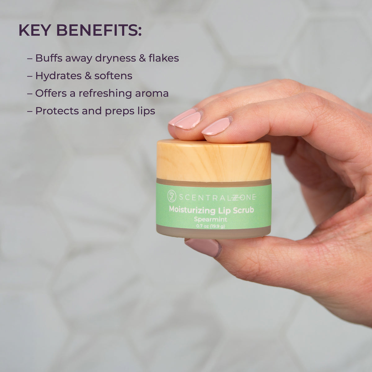 A hand is holding Scental Zone .7oz Moisturizing Lip Scrub in Spearmint in a jar with a light green wrapper and bamboo lid. Displayed next to the jar is text displaying the key benefits. 