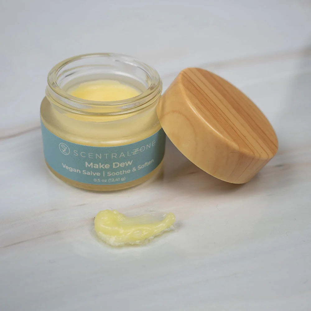 Open jar of Scentral Zone Make Dew Vegan Salve with a dollop of the product to show the texture and consistency of the product. The salve is in a blue wrapped container with a wooden lid labeled to smooth and soften. 
