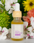 Scentral Zone 0.5 fl oz Velvet Face Serum, a yellow rejuvenating serum with cucumber and seaweed, in a glass dropper bottle with a wooden collar and white dropper top. Product is displayed in front of various plants and flowers as the background. 